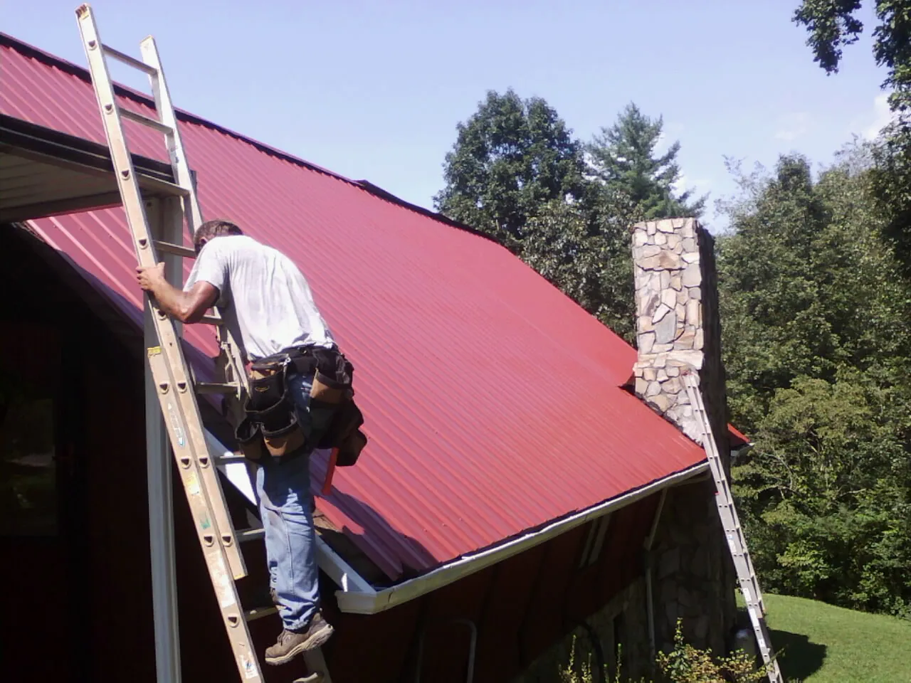 Roofer on a ladder beside a red metal roof and stone chimney — Soffit & Fascia Repair by Roofing Brothers in Hadlyme